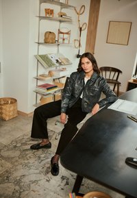 Zwart leren cropped jack met voorzakken, witte shirt, zwarte wijde broek en glanzende zwarte loafers. Interieur met lichte decoratie.