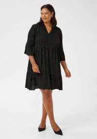 Vestido negro con escote en V, mangas tres cuartos, diseño escalonado y rayas metálicas verticales. Textura suave, a la altura de la rodilla, combinado con zapatos negros.