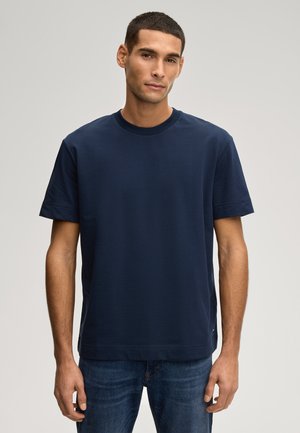 Homme debout portant un t-shirt uni bleu foncé à manches courtes et un jean bleu sur un fond gris clair.