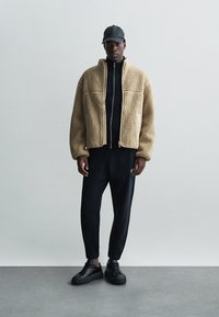 Teddy fleece jacket in tan with zip closure, black fitted top, black sweatpants, and black sneakers. Accessories include a black cap.