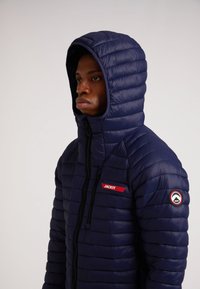 Marineblå pufferjakke med quiltet design, høj krave, lynlås foran og mærkning på brystet og ærmet. Materialet ser glansfuldt ud.