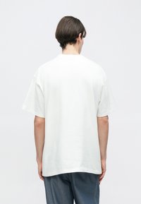 Λευκό βαμβακερό μπλουζάκι με άνετη εφαρμογή, κοντά μανίκια και στρογγυλεμένο λαιμό, εμφανιζόμενο από πίσω. Λείο ύφασμα, μίνιμαλ σχέδιο.