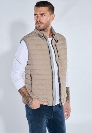 Beige quilted vest with a high collar, front zipper, and side pockets, paired with a white long-sleeve shirt and dark jeans.