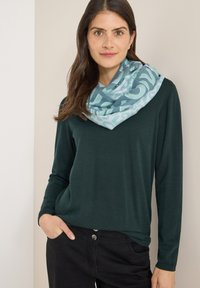Haut à manches longues vert foncé associé à une écharpe bleu clair à motifs abstraits et texture douce, portée avec un pantalon noir.