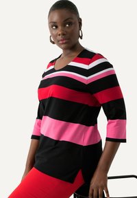 Frau mit Creolen, die ein gestreiftes Oberteil in Schwarz, Rot, Pink und Weiß sowie rote Hosen trägt, sitzt mit einer Hand, die auf einem schwarzen Stuhl ruht.