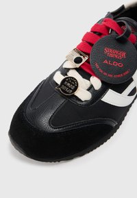 Fekete sportcipő piros és fehér részletekkel, texturált anyagokból, kerek címkével, valamint „ÜDVÖZÖLJÜK AZ FENTI OLDALON” és „IDEGEN DOLGOK” feliratú gombokkal.