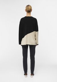 Maglione oversize nero e beige lavorato a maglia con un design a blocchi di colore diagonali. Il modello indossa jeans scuri e sandali semplici.
