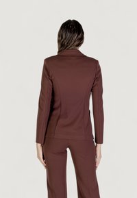 Hnědý tailored blazer vyrobený z elastického materiálu. Má strukturovaný límec a přiléhavý střih se švy po stranách a dlouhými rukávy.