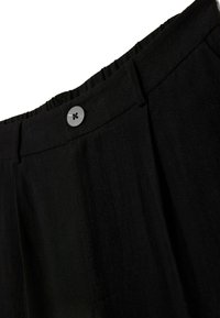 Gros plan sur la ceinture d’un pantalon noir avec une fermeture par bouton et des passants de ceinture, montrant la texture du tissu et une bande élastique à l’arrière.