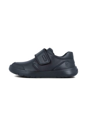 Zapato sintético negro con punta redonda, correa de Velcro y suela texturizada. Presenta un ligero tacón y sin patrones o marcas visibles.