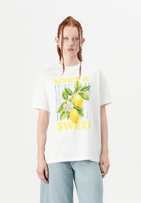T-shirt bianco in cotone con una grafica di limone giallo, foglie verdi e il testo "SOUR & SWEET" in grassetto giallo. Vestibilità rilassata, maniche corte.