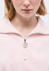 Νεαρή γυναίκα με κοντά ξανθά μαλλιά που φοράει ροζ ανοιχτόχρωμο τοπ με φερμουάρ και κυκλικό μοτίβο στο τρέμουλο του φερμουάρ, καθώς και χρυσούς κρίκους σκουλαρίκια.