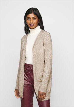 Beige neulecardigan, jossa on avoin etuosa, pitkät hihat ja ribbireunat. Päällä valkoinen poolopaita ja burgundinväriset tekonahkahousut.