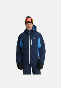 Marineblaue Skijacke mit hellblauen Akzenten, ausgestattet mit einem Reißverschluss vorne, Kapuze und mehreren Taschen. Vollendet mit einem Spinnenlogo auf der Brust.