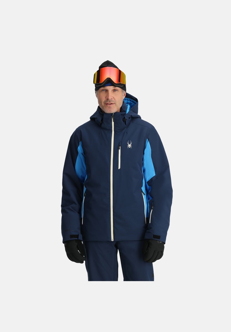 Marineblaue Skijacke mit hellblauen Akzenten, ausgestattet mit einem Reißverschluss vorne, Kapuze und mehreren Taschen. Vollendet mit einem Spinnenlogo auf der Brust.