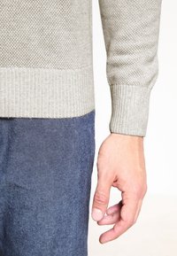 Pull texturé gris clair avec un ourlet côtelé, associé à un pantalon en denim foncé. La main visible est détendue contre le tissu.