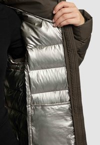 Puffjacke med en metallisk sølvforing, der har et mørk olivenfarvet ydre og en quiltet struktur. Lynlåslomme synlig.