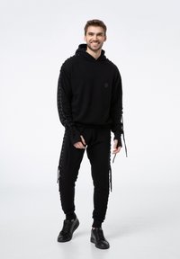 Czarna bluza z kapturem z detalami na rękawach, połączona z dopasowanymi spodniami dresowymi. Model w czarnych sneakersach, stojący na neutralnym tle.