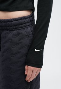 Crna majica dugih rukava s prilagođenim manžetama, u kombinaciji s tamnosivim treniranim hlačama s teksturiranim uzorkom i elastičnom uključnicom. Nikeov logo na majici.