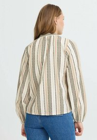 Kvinde med langt hår if�ørt en cremfavet langærmet bluse med lodrette sorte og orange striber samt blå jeans, der vender væk.