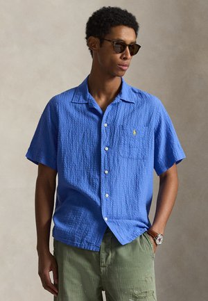 Muž nosiaci modrú textúrovanú košeľu s krátkym rukávom s gombíkmi, zelené roztrhané nohavice, slnečné okuliare a náramkové hodinky, pózujúci na jednofarebnom pozadí.