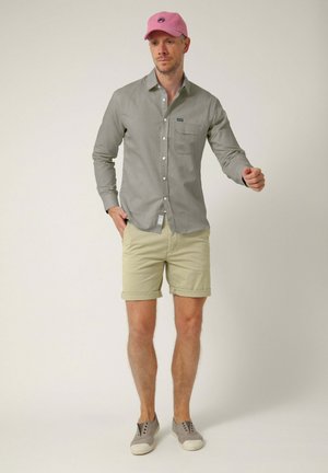 Hombre con gorra rosa, camisa gris de manga larga con botones, pantalones cortos beige y zapatos grises sin cordones, de pie con una mano en el bolsillo.