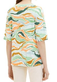 Femme portant un haut ample à manches courtes avec un motif abstrait ondulé en vert, orange, blanc et moutarde, associé à un pantalon clair.