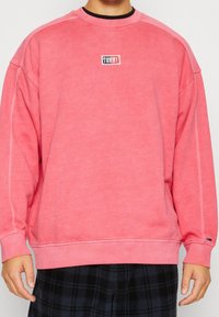 Sudadera rosa con cuello redondo, puños y dobladillo acanalados. Presenta un pequeño logo "TOMMY" en negro y rojo. Tejido suave y texturizado.