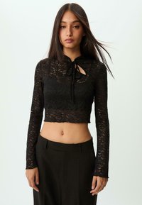 Cropped top in pizzo nero con maniche lunghe, scollo a chiave con fiocco, dettagli testurizzati e orlo a scallop. Abbinato a pantaloni scuri a vita alta.