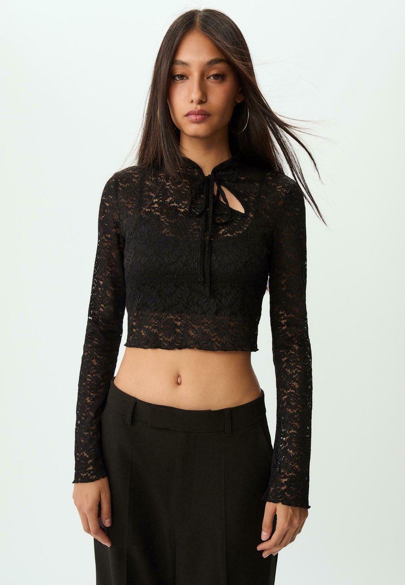 Cropped top in pizzo nero con maniche lunghe, scollo a chiave con fiocco, dettagli testurizzati e orlo a scallop. Abbinato a pantaloni scuri a vita alta.