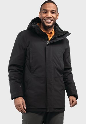 Lächelnder Mann, der eine schwarze Kapuzen-Winterjacke über einem orangefarbenen Hemd trägt, stehend vor einem schlichten hellen Hintergrund.