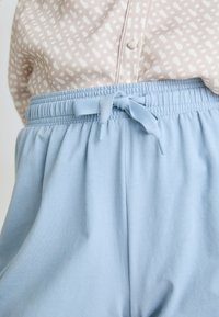 Lichtblauwe elastische taillebandbroek met een knoopdetail aan de voorkant, gemaakt van zacht materiaal, gecombineerd met een geblokte beige top.