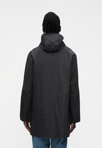 Černá deštná bunda s pevnou kapucí, vyrobená z mačkaného materiálu. Má dlouhé rukávy a volný střih, sahající pod boky.