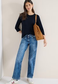 Haut bleu marine à manches trois-quarts, jeans à jambes larges bleu clair avec fermeture à boutons, et sac à épaule en daim beige. Baskets blanches avec accents rouges.