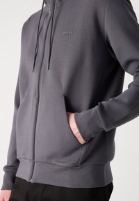 Sudadera con cremallera en tejido gris oscuro, con un sutil logo "BOSS", cordones ajustables y bolsillos laterales para mayor comodidad.
