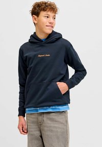 Sweat à capuche bleu marine en tissu doux, doté d'une poche kangourou et d'un texte brodé en or ; superposé à une chemise à rayures bleues.