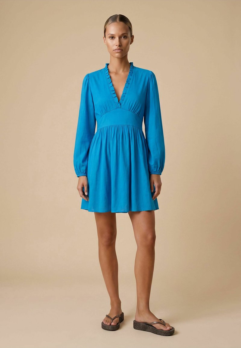 Femme debout face à l’avant, vêtue d’une robe bleu vif à manches longues avec un décolleté en V profond et un col à volants, associée à des sandales compensées marron.