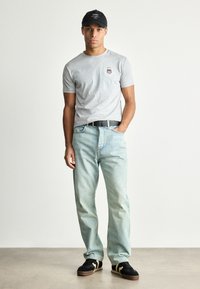 Grå kortærmet t-shirt med rund halsudskæring og logo, blå løstsiddende jeans, sort kasket og sorte sko med gule detaljer.