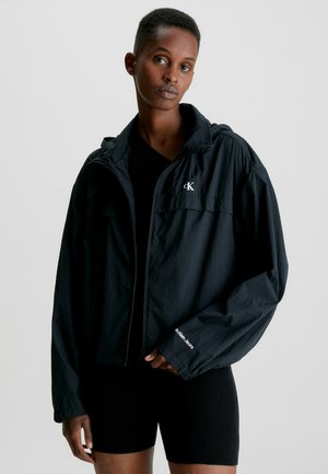 Chaqueta ligera negra con capucha, cierre de cremallera y logo bordado en el pecho. Lleva una camiseta negra ajustada y pantalones cortos.