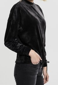 Top de terciopelo negro de manga larga con cuello alto. Presenta un cordón en el dobladillo y una textura suave y lisa. Combinado con jeans oscuros.