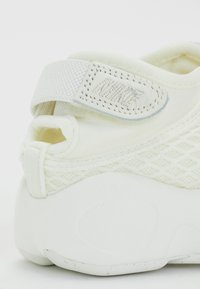 White sneaker with mesh upper, curved heel, and padded strap. Features textured accents and a stitched logo on the strap.