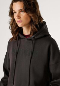 Felpa nera con cappuccio dotata di coulisse, con la scritta "PEPE JEANS" in perline iridescenti sul petto, dalla finitura morbida e strutturata.