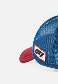 Niebieska siatkowa czapka z czerwoną daszkiem, z białą łatą z czarnym logo. Tekstura zawiera przewiewny design oraz solidne akcenty.