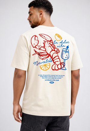 Man wearing cream T-shirt with red lobster, lemon slices, cocktail glass, and Italian text "la dolce vita" printed on back.