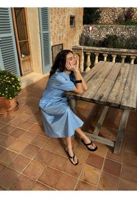 Žena u plavoj haljini sjedi na popločanom dvorištu, naslanjajući glavu na ruke za drvenim stolom blizu kamenog zida i vrata s griljama.