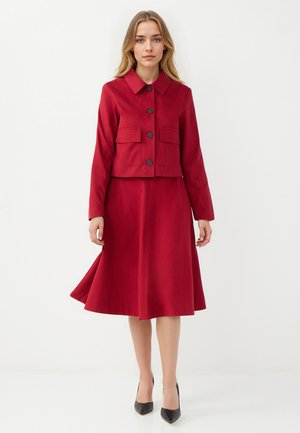Ensemble de veste et robe rouges, comprenant une veste à col avec deux poches et des boutons noirs, accompagnée d'une jupe évasée. Talons hauts noirs.