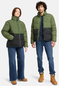 Chaquetas de plumas verdes y negras con cuellos altos, que presentan bolsillos frontales y cremalleras en contraste, combinadas con vaqueros azules y botas marrones.