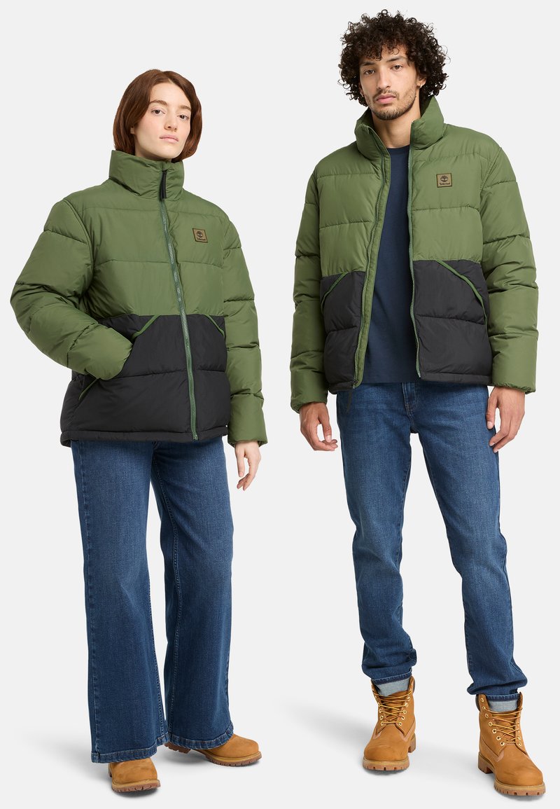 Chaquetas de plumas verdes y negras con cuellos altos, que presentan bolsillos frontales y cremalleras en contraste, combinadas con vaqueros azules y botas marrones.