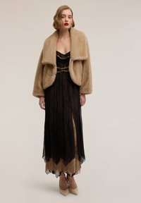 Giacca in pelliccia ecologica beige sopra un abito nero di pizzo con dettagli dorati, caratterizzato da un corpetto aderente e una gonna fluente. Scarpe con tacco a punta beige.