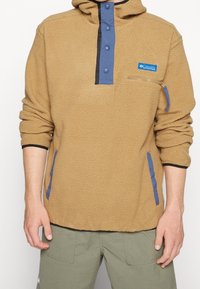 Pullover in pile color sabbia con zip a un quarto, bottoni blu e tasche laterali. Presenta una tasca frontale sul petto con dettaglio a zipper. Tessuto testurizzato.